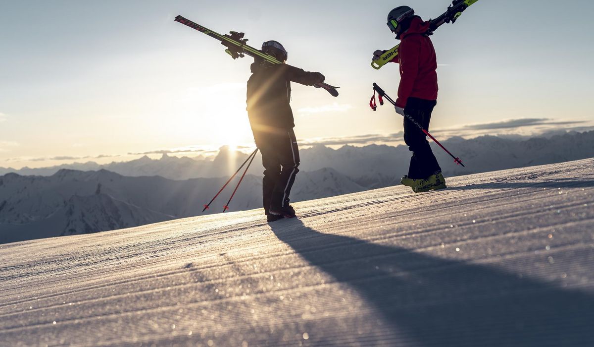 Zwei Skifahrer mit Skiern auf den Schultern stehen auf einem Berggipfel. Die Sonne geht im Hintergrund unter, und ihr Schatten fällt auf den Schnee.