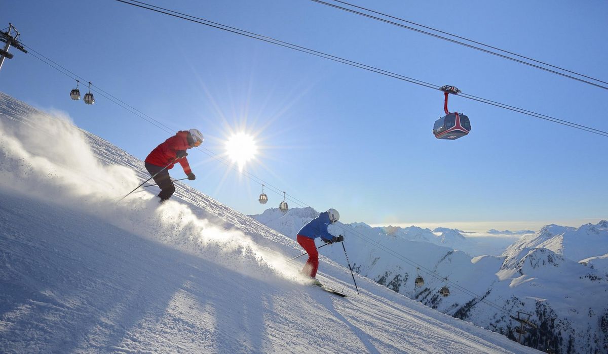 Zwei Skifahrer in roter und blauer Kleidung fahren einen sonnigen, schneebedeckten Hang hinunter. Skilifte und schneebedeckte Berge sind im Hintergrund sichtbar.