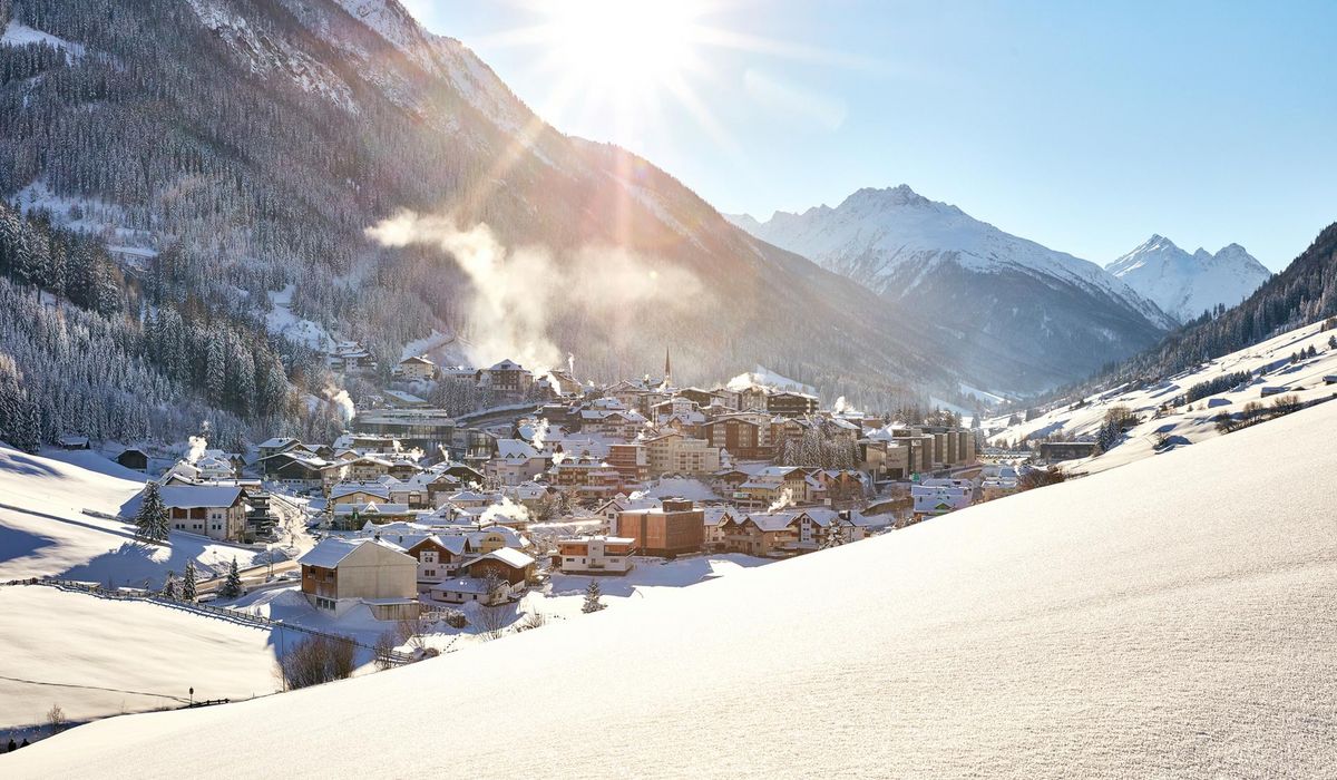Die Ortschaft Ischgl in einem verschneiten Tal, umgeben von schneebedeckten Bergen. Die Sonne strahlt hell am blauen Himmel.