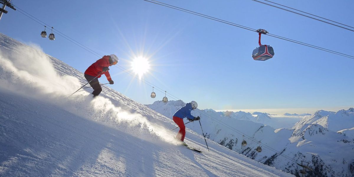 Zwei Skifahrer in roter und blauer Kleidung fahren einen sonnigen, schneebedeckten Hang hinunter. Skilifte und schneebedeckte Berge sind im Hintergrund sichtbar.