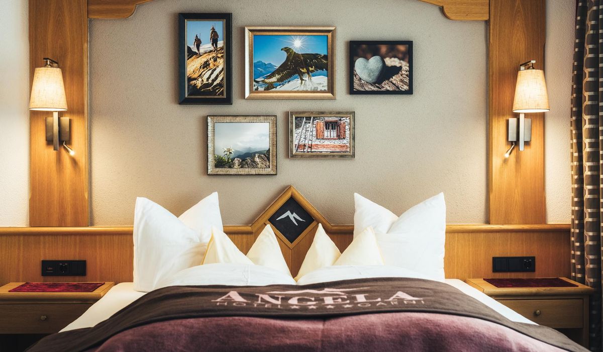 Ein Hotelzimmer mit einem Doppelbett. Über dem Bett hängen gerahmte Bilder von Berglandschaften, einem Adler und einem Herz aus Stein.