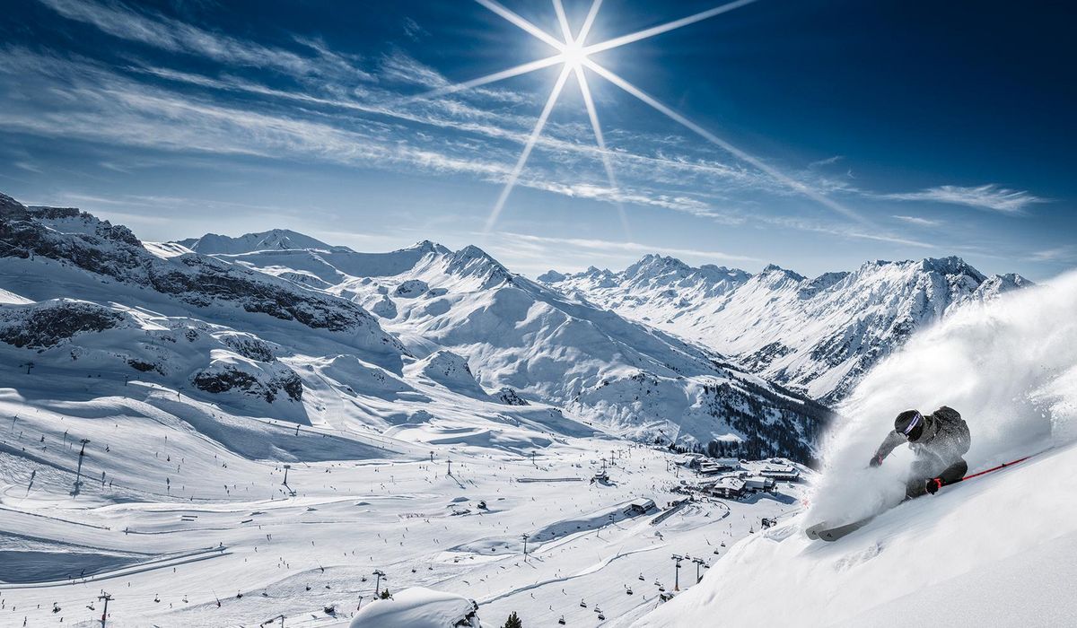 Skifahrer fährt durch tiefen Schnee an steilem Hang, weite verschneite Berglandschaft im Hintergrund.