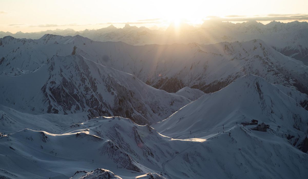 Eine weite Ansicht auf schneebedeckte Berggipfel, die im Sonnenlicht liegen. Am Horizont strahlt die Sonne und erhellt die Landschaft.