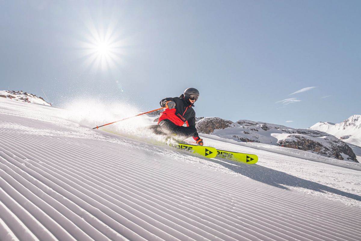 Skifahrer in Kurve auf frisch präparierter Piste, Schneestaub sprüht unter klarem Himmel.