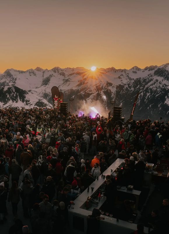 Menschenmenge vor Bühne auf Bergterrasse bei Sonnenuntergang, umgeben von verschneiten Gipfeln.