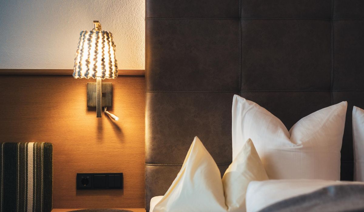 Ein Bett in einem Hotelzimmer mit einem braunen, gesteppten Kopfteil und weißen, dekorativen Kissen. Eine moderne Wandlampe mit geflochtenem Schirm hängt darüber.