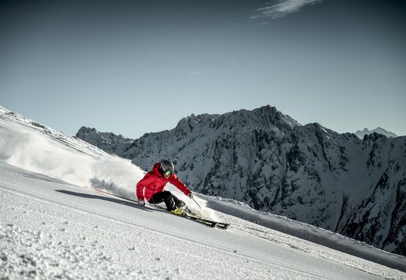 Ein Skifahrer in roter Jacke und schwarzer Hose fährt schnell einen schneebedeckten Berghang hinunter, wirbelt Schnee auf und ist in einer tiefen Kurve.